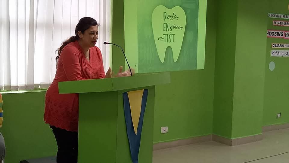 Dr. Nisha Bali speaking at a podium beneath a 'Doctor Engineer Artist' wall during a school career and health talk